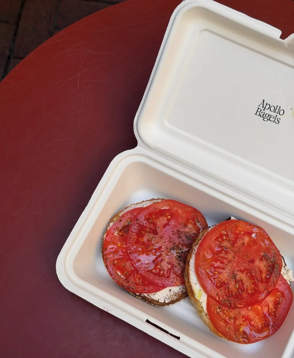 An Apollo Bagels tomato and cream cheese bagel open in its takeout box on a red table