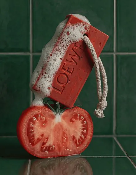 A red Loewe tomato-shaped soap bar resting on a sliced tomato against green tiles